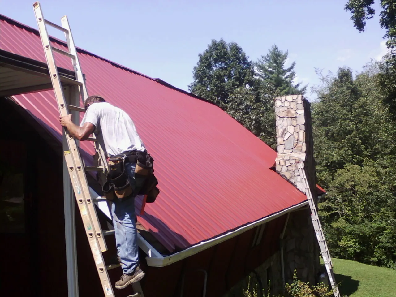 Roofer on a ladder beside a red metal roof and stone chimney — Soffit & Fascia Repair by Roofing Brothers in Owatonna